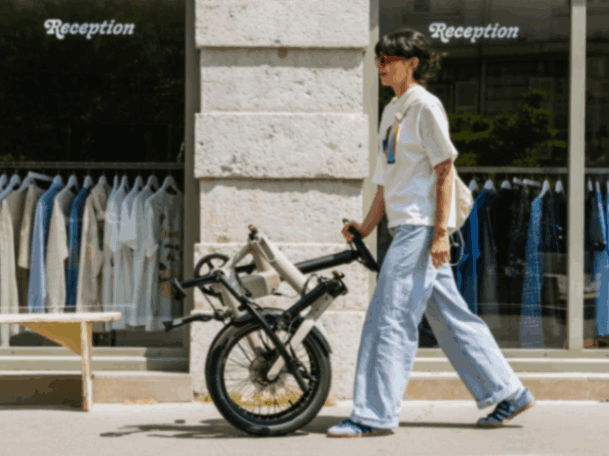 Casually dressed woman pushing a folded electric bike past a city clothes shop